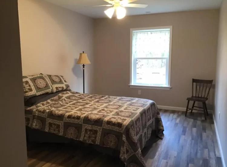 A modestly decorated guest bedroom with a patterned bedspread, wooden flooring, and a single chair, creating a comfortable, minimalist space.