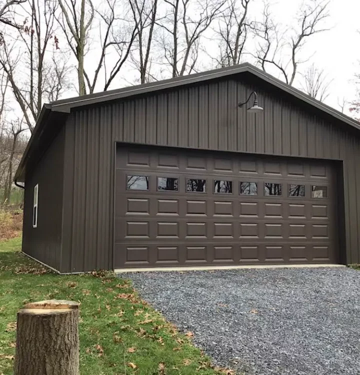 A standalone garage with a dark brown exterior and large garage door, located in a wooded area with a gravel driveway.