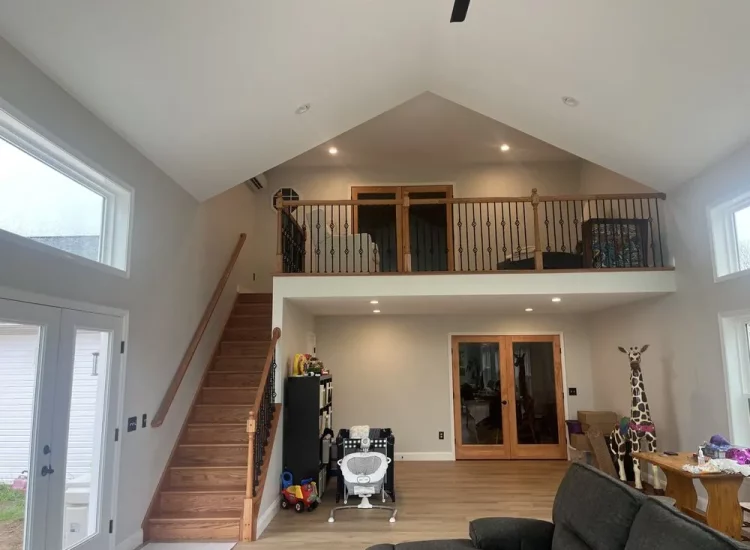 An open and airy living room featuring high ceilings, a charming loft balcony with wooden railings, and large windows that allow ample natural light, creating a spacious and welcoming environment.