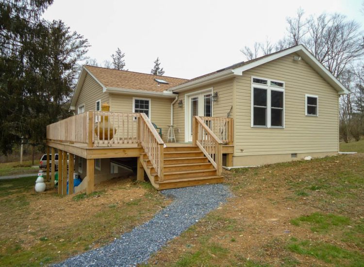 Newly built beige home addition with a spacious wooden deck and stairs, providing a comfortable outdoor living space in a natural setting.