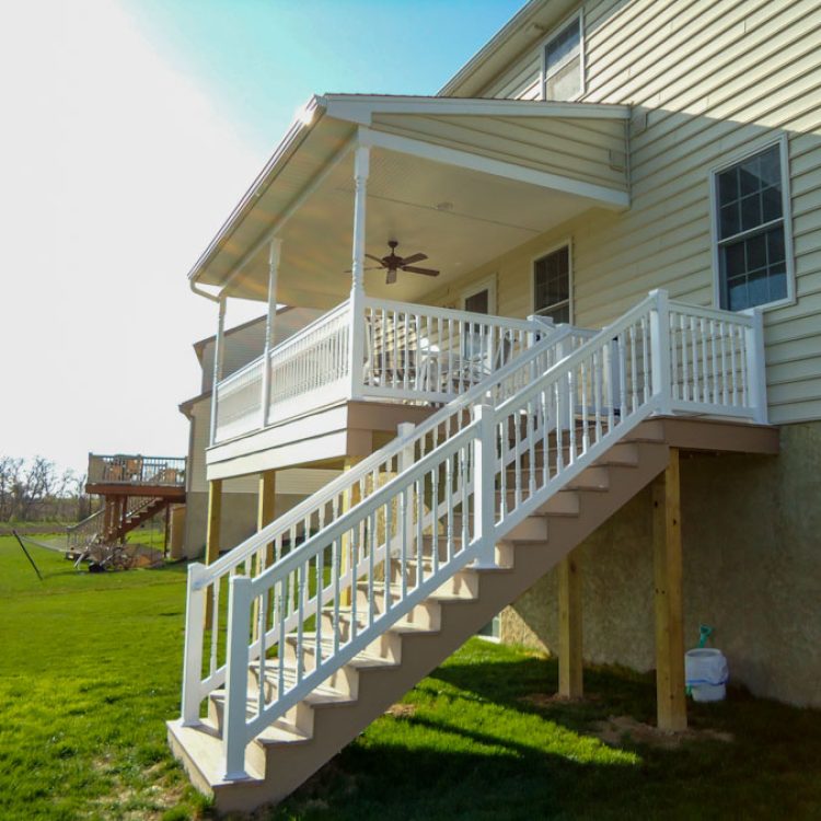 A stylish elevated porch with white railings and an outdoor ceiling fan, providing a comfortable and airy outdoor living space adjacent to a suburban home.